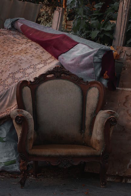 Inside a home, an ornate vintage armchair with carved wooden details and upholstery in muted tones is positioned in front of a partially covered window or doorway. On the left side of the image, a patterned rug or blanket drapes over a piece of furniture or surface, with a rolled-up quilt or fabric with fabric cover resting on top. To the right, part of a wooden structure or shelving unit is visible, supporting cardboard and plastic-wrapped items, suggesting packing or decluttering activities associated with house removals. The environment has natural lighting, likely from an open door or window, indicating a moving or packing process. This scene reflects elements involved in home relocation, furniture transport, or packing and moving services, with careful handling of furnishings and household items as characteristic of professional removals. Man with Van Lower Morden offers handling of such household items during professional moves.