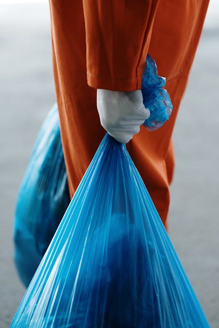 The image shows a person wearing an orange protective jumpsuit and a white glove, holding a large blue plastic rubbish bag by its gathered top handle. The individual is standing outdoors on a paved surface, with a blurred background suggesting an open space or driveway. The scene likely relates to waste collection, disposal, or cleaning activities associated with home decluttering or removal services. This visual context aligns with furniture or waste removal processes, consistent with professional removals or packing and moving tasks offered by Man with Van Lower Morden. The image emphasizes the action of handling waste materials in preparation for a house move or clearance, highlighting the importance of proper waste management during relocation projects.