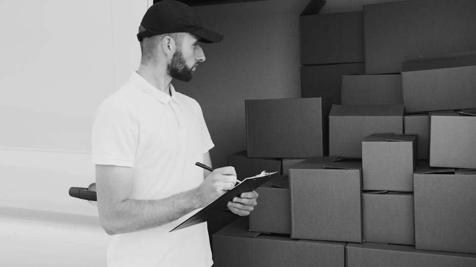 A man wearing a black cap and white polo shirt is standing indoors, holding a clipboard and pen while inspecting and taking notes on a piece of paper. Behind him, there are several stacked cardboard b