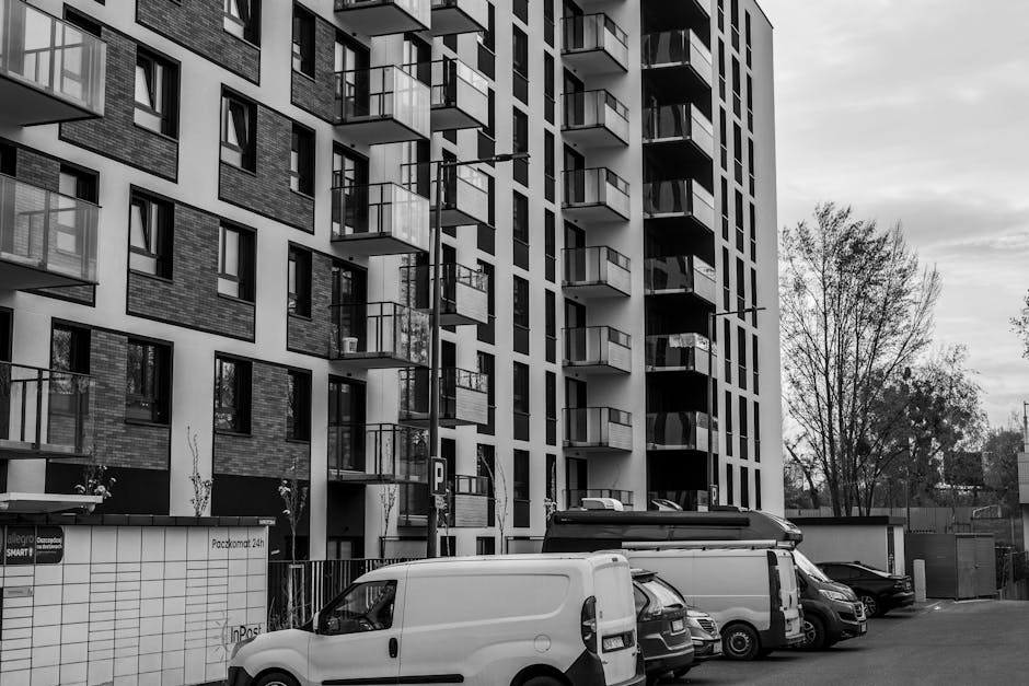 Black and white photograph of a modern multi-storey apartment building with multiple balconies featuring glass railings, situated along a street. In front of the building, a parking area contains seve