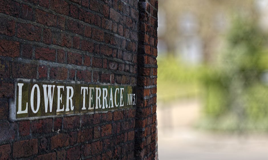 Close-up view of a brick wall with a street sign reading 'Lower Terrace NW3' attached to it. The sign is made of metal with white lettering on a green background. In the background, there is a partially visible open doorway leading inside the house, showing a glimpse of a cluttered interior with cardboard boxes, plastic wrap, and packing materials placed on the floor near the entrance. To the right, an area of pavement and greenery is visible, with soft natural lighting illuminating the scene. This image captures elements related to home relocation and the packing and moving process, including the preparation of belongings and the logistics involved in furniture transport, as seen in the placement of boxes and wrapping materials near the doorway, often managed by professional removals services like Man with Van Lower Morden.