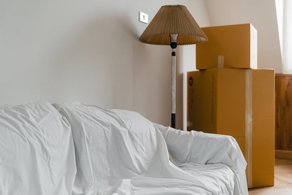 Interior scene showing a living room area with a white sofa draped in a white sheet, positioned against a plain beige wall. To the right, there are three cardboard moving boxes stacked near a wooden piece of furniture. A tall, standing lamp with a brown lampshade is situated between the sofa and the boxes, casting soft light into the room. The boxes are sealed with packing tape, indicating they are prepared for a home relocation process. The environment suggests a move-in or move-out situation, with the packing and furniture arranged for transportation. The scene highlights the typical elements of furniture transport, packing materials, and moving logistics often involved in house removals managed by services like Man with Van Lower Morden, supporting efficient and careful furniture transport during house moves.