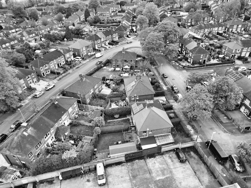 An aerial black-and-white photograph of a residential area showing a mix of detached and semi-detached houses with pitched roofs, surrounded by trees and small gardens. The scene includes narrow streets with parked cars on both sides, and some vehicles are moving through the area. In the foreground, there are backyard gardens with fences, sheds, and gardening equipment. A large tree dominates the right side of the image, with branches extending over the adjacent houses. The area appears quiet and well-organized, with houses closely spaced but each having its own distinct outdoor space. This view captures the typical suburban layout suitable for home relocation or furniture transport purposes, as part of managing house removals within Merton, supported by Man with Van Lower Morden.