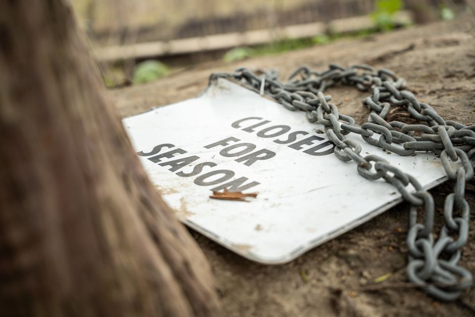 A weathered white rectangular metal sign lying on the ground in an outdoor environment, partially obscured by a large tree trunk in the foreground. The sign has bold black text that reads 'CLOSED FOR SEASON,' and is secured with a heavy metal chain looped around it, suggesting it is meant to restrict access. The sign appears dirty and slightly worn, with some dirt and small leaves on its surface, indicating it has been exposed to the elements for some time. Behind the sign, there is a dirt surface with a wooden fence or structure in the background, and the lighting is natural with soft shadows, consistent with daytime outdoor conditions. The scene implies a setting such as a park, outdoor facility, or garden that is temporarily unavailable, which could relate to house removals or property management in the context of relocation services provided by Man with Van Lower Morden.