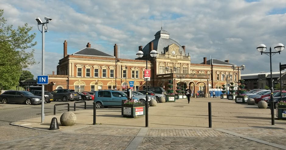 A wide view of Morden Station's outdoor parking area under a partly cloudy sky, with several parked cars of various sizes and colours situated in front of the station building. The building features traditional brick architecture with large windows, a central clock, and decorative elements on its façade. The foreground includes a paved pedestrian area with black bollards, metal railings, and large spherical stone bollards positioned near flower planters with hanging baskets. A few people are visible walking near the entrance, and a tall street lamp with multiple lights stands to the left. The scene captures a typical day of the station's vicinity, which is relevant for house removals and packing and moving activities conducted by Man with Van Lower Morden, including the loading process of furniture and boxes into vehicles for home relocation services.