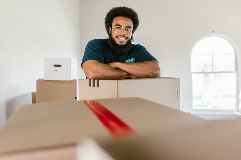 A man with dark curly hair and a beard is smiling while leaning on a stack of cardboard boxes inside a well-lit room with a large arched window in the background. The boxes vary in size, with some wrapped in plastic or packing tape, and are arranged on the floor in front of him, suggesting the process of packing for a home relocation. The scene includes other packing materials such as smaller boxes and a white storage box on top of one of the larger boxes. The natural light coming through the window illuminates the space, creating a clean and organized atmosphere typical of professional removals. The setting indicates an ongoing packing and furniture transport phase, and the overall environment reflects a professional moving service like Man with Van Lower Morden, supporting efficient home removals and loading activities.