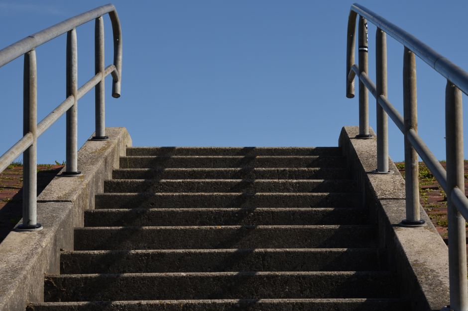 A set of outdoor concrete stairs leading up to a higher level with metal handrails on both sides, casting shadows on the steps under bright sunlight against a clear blue sky. The stairs appear to be part of a residential or public property, possibly used during a home relocation or furniture transport process. The surrounding environment includes portions of brick paving or pathway at the base of the stairs, and the area is well-lit, emphasizing the structural details of the steps and railings. This image exemplifies the logistical considerations involved in navigating narrow staircases within house removals and the need for careful planning during packing and moving activities, as handled by companies like Man with Van Lower Morden.