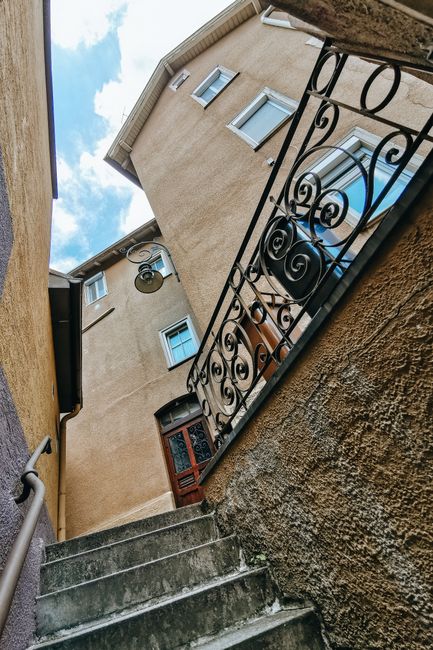 A narrow outdoor staircase leading up to an apartment entrance on the upper floor of a residential building on Lower Morden Estates. The stairs are made of concrete with visible wear and patches of moss, bordered by a textured brown wall on the right and a smooth yellow wall on the left. A black wrought iron railing with decorative scrollwork runs along the right side of the staircase for safety. At the top of the stairs is a small landing with a wooden door featuring a glass panel and a brass handle, positioned beneath three small windows on the beige exterior wall of the multi-story building. The sky above is partly cloudy, providing natural daylight to the scene. This image exemplifies the typical exterior features involved in home relocations or furniture transport logistics to and from upper-level residences, as handled by Man with Van Lower Morden in their house removal services.