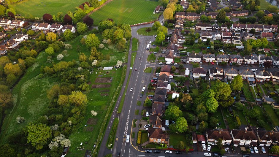 Aerial view of a residential neighbourhood featuring a main road with moving traffic, lined by detached and semi-detached houses with gardens, trees, and driveways. To the left, there is a green space with grass, bushes, and flowering trees, possibly a park or community garden. The houses have varied roofing and are surrounded by hedges and lawns. Trees with fresh foliage are visible throughout the area, indicating spring or early summer. The scene includes parked cars along the street and within driveways, with some vehicles potentially used by local removal services. This landscape captures the calm suburban environment where house removals and furniture transport are commonly coordinated by companies like Man with Van Lower Morden, providing an overview of the typical setting for home relocation tasks regulated by local traffic and access considerations.