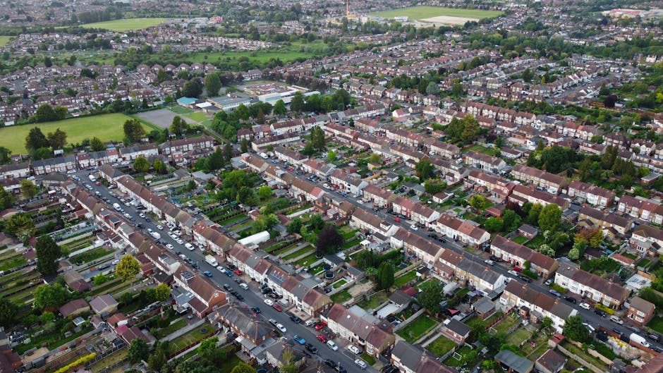 An aerial view of a suburban residential area showing rows of terraced houses with small, well-maintained gardens, some equipped with garden sheds and outdoor furniture. Driveways and parking areas are filled with various cars parked beside the houses. The street runs through the neighborhood, with trees and greenery scattered throughout, providing a sense of community and comfort. In the foreground, a large commercial building with a parking lot is visible, adjacent to the houses. The image captures a clear day with natural daylight, highlighting the typical environment where house removals and furniture transport services by Man with Van Lower Morden could be involved in a home relocation process, including packing, loading, and transporting household items using vans and equipment visible on-site.