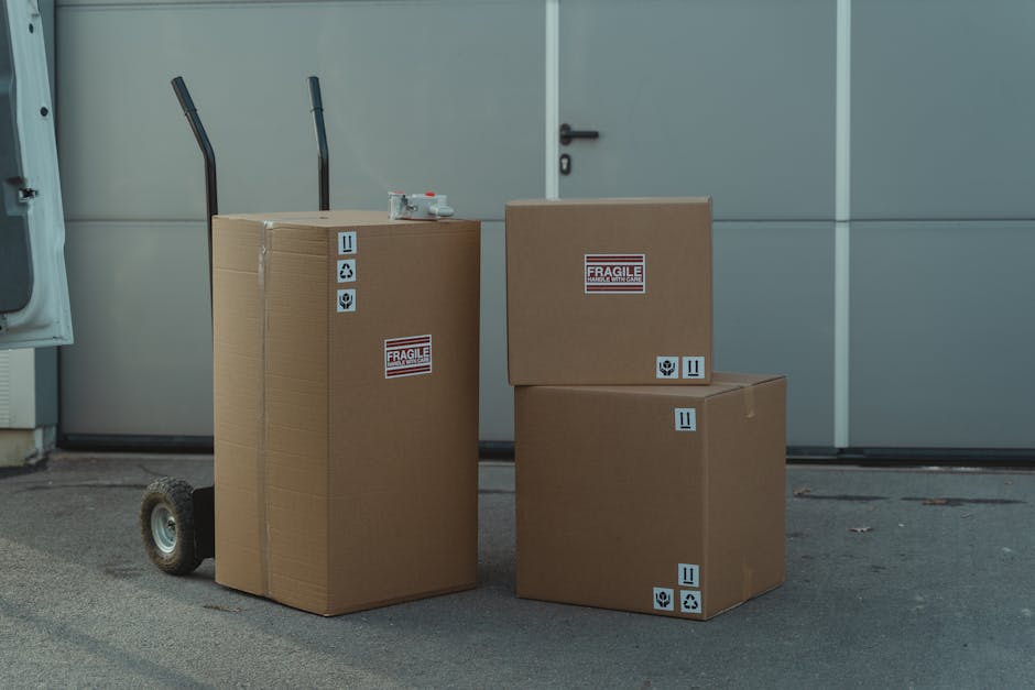 Four cardboard boxes of varying sizes are stacked and placed on the pavement outside a building, with some boxes positioned on a small, wheeled trolley. The boxes are sealed with packaging tape and display labels indicating fragility, handling instructions, and recycling symbols. Two tall, black hand trucks with ergonomic handles are situated behind the boxes, suggesting preparation for moving or furniture transport. The background features a large, light grey industrial-style door with a black handle and a white wall, illuminated by natural daylight. This scene is associated with home relocation or packing and moving services provided by Man with Van Lower Morden, highlighting packing, logistics, and loading processes typical of same-day removal operations.