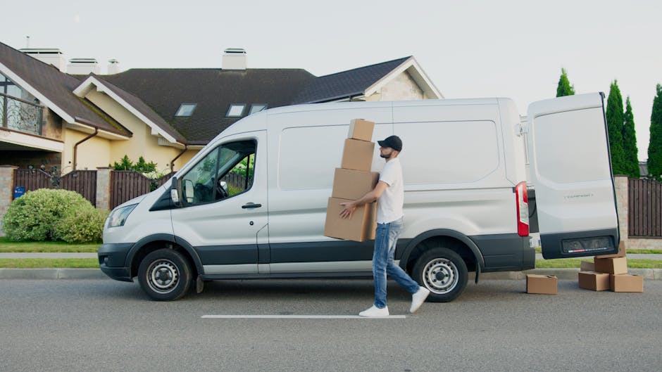 A man wearing casual clothing and a cap is outside on the pavement, carrying three cardboard boxes stacked together towards a white cargo van parked on the street in a residential area. The van has its side door open, revealing an interior prepared for a home relocation. Several more cardboard boxes are placed on the ground near the open rear door of the van. The background features modern houses with pitched roofs, chimneys, and well-maintained gardens, including bushes and tall trees. The scene is illuminated with natural daylight, indicating daytime. This image depicts a house removal process involving furniture transport and packing, as part of a professional moving service by Man with Van Lower Morden, supporting efficient loading and home relocation logistics.